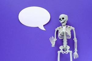 Skeleton on violet background with white blank paper ellipse speech bubble. Anatomical plastic model human skeleton with round text balloon. Empty dialog cloud, mockup, copy space. Purple Halloween. photo