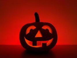 Halloween holiday background with traditional smiling jack-o-lantern. Big carved glowing pumpkin stands on the surface in dark night. The back wall highlighted in red. photo