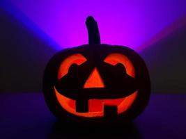 Halloween holiday purple background with orange smiling jack-o-lantern. Big carved glowing pumpkin stands centered on table in dark night. Back wall highlighted in violet triangle over pumpkin. photo