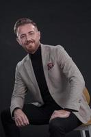 Portrait of adult businessman wearing trendy suit and sitting in modern studio on stylish chair against the black background photo