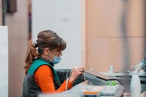 a woman sitting on a chair and assembles a screen for a machine, wearing a protective mask on her face due to a coronavirus pandemic photo