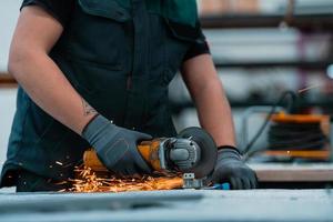 interior de la fábrica de ingeniería de la industria pesada con un trabajador industrial que usa una amoladora angular y corta un tubo de metal. Lleva una máscara en la cara debido a la pandemia del coronavirus foto