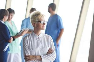 female doctor with glasses and blonde hairstyle standing in front of team photo