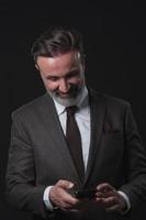 Chat with client. Good looking young man in full suit using his smart phone and smiling while standing against grey background. photo