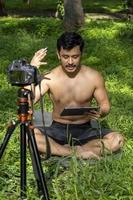 Young man practicing yoga asana, balance, meditating while standing on one leg on sports mat on green grass in park. Using tablet for online class. photo