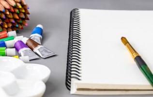 cuaderno de bocetos de acuarela en blanco sobre fondo gris con paleta de cerámica de pincel, tubo y lápices de colores foto