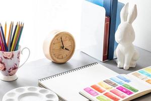 espacio de trabajo del artista de la acuarela con cuaderno de bocetos en blanco, paleta, tubo de color, lápices de colores, reloj de madera y estatua de conejo blanco, vista lateral sobre fondo gris. foto