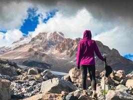 Back view inspirational view woman hiker stand on rocky terrain viewpoint look to Kazbek mountain peak in KAzbegi national park. Gergeti glacier KAzbek trekking trail route photo