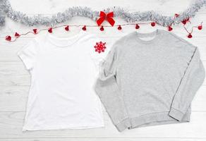 cierre la camiseta blanca con plantilla en blanco, la sudadera con capucha gris con espacio de copia y el concepto de vacaciones de navidad. camiseta de maqueta de vista superior, lazo rojo sobre fondo de madera blanca. feliz año nuevo decoraciones foto