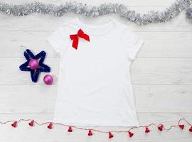 Close up white blank template t shirt with copy space and Christmas Holiday concept. Top view mockup t-shirt and red bow on white wooden background. Happy New Year decorations accessories. Xmas outfit photo