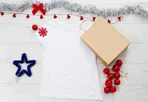 Close up white blank template t shirt with copy space paper bag. Christmas Holiday concept. Top view mockup t-shirt, star, white wooden background. Happy New Year decorations accessories. Xmas outfit photo