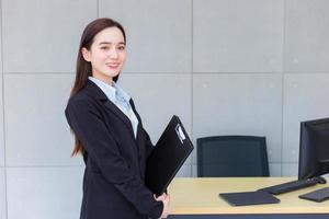 mujer joven de negocios profesional asiática en traje negro sonríe felizmente con confianza y mira la cámara mientras trabaja y sostiene el portapapeles en la oficina. foto