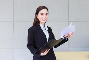 Professional Asian smart business woman manager is smiling confidently while she holds and opens document clipboard in her hand at work office. photo