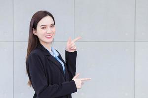 Una mujer trabajadora asiática hermosa y profesional que usa traje negro está señalando con la mano para presentar algo en la pared de la oficina. foto