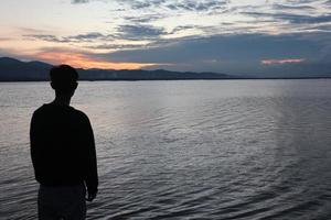 silueta de un joven parado junto al lago disfrutando de la puesta de sol. ambiente tranquilo en la naturaleza foto