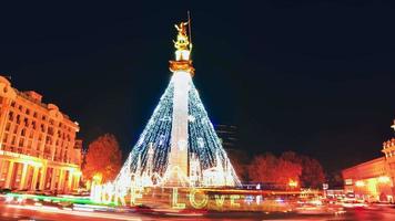 reveladores coches de lapso de tiempo pasan en la rotonda dejando senderos de luz con un árbol de Navidad decorado en la plaza de la libertad y turistas tomando fotografías