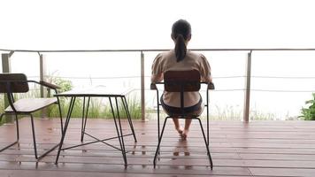achterzijde visie van vrouw zittend Aan terras en op zoek Bij zee van mist in ochtend- video