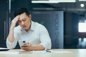 hombre de negocios asiático molesto que trabaja en la oficina, leyendo malas noticias por teléfono, hombre deprimido en el trabajo foto