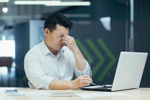 hombre de negocios asiático cansado que trabaja hasta tarde en la oficina, dolor de ojos, hombre con laptop. foto