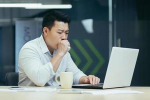 Sick at work. A young Asian businessman keeps coughing, covers his mouth at the desk in the office photo