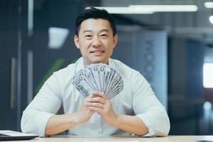 A happy young Asian businessman holds a cash of money in his hands and waves it, sitting in a office photo