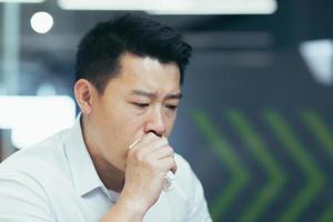 Photo close-up portrait of an Asian man in a shirt, a sick businessman coughs into a tissue