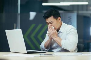 Sick man in office coughs and runny nose, Asian businessman with cold works in office on laptop photo