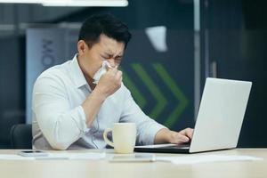 hombre enfermo en la oficina tose y moquea, hombre de negocios asiático con trabajos en frío en la oficina en la computadora portátil foto