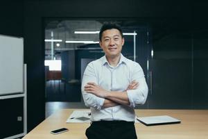 retrato de un exitoso jefe asiático en una oficina moderna, hombre de pie con los brazos cruzados foto