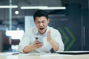 hombre de negocios enojado en la oficina gritando por teléfono, hombre asiático frustrado con el correo electrónico recibido foto