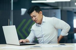 hombre de negocios asiático que trabaja en la oficina, con dolor de espalda severo, trabajador cansado que trabaja con una computadora portátil foto