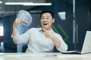 A happy young Asian businessman holds a cash of money in his hands and waves it, sitting in a office photo