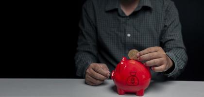 A man's hand is dropping digital gold coins into a red piggy bank on a table inside a dark room with black walls and empty spaces. photo