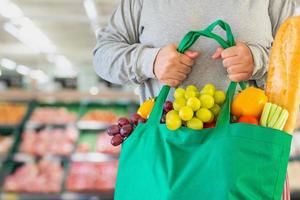 Customer hold reusable green shopping bag with fruit and vegetable over Supermarket aisle blur defocused product shelves interior bokeh light background photo