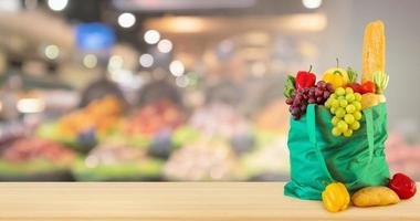 Green shopping bag with groceries product on wood table top with abstract blur colorful Fruits in display basket in supermarket grocery store defocused background with bokeh light photo