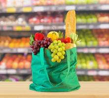 Fresh fruits and vegetables in reusable green shopping bag on wood table top with supermarket grocery store blurred defocused background with bokeh light photo