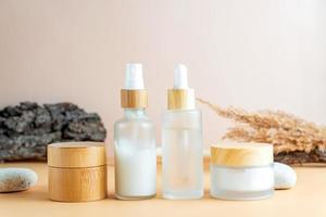 White frosted glass cosmetics bottles and jars with pamboo lids with tree bark, pampas grass, stones on beige background. Autumn fall beauty products set. photo