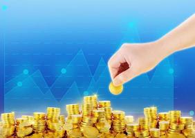 one hand is putting gold coins on pile of gold coins with graphic blue background photo