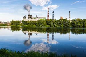 tuberías de aserradero de planta empresarial de carpintería con un hermoso reflejo en el agua azul del río. concepto de contaminación del aire. paisaje industrial contaminación ambiental residuos de central térmica foto