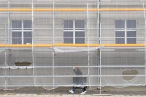 Detail shot of a facade of a house in the city. There is scaffolding in front of the facade. A net is stretched over the scaffolding. Two women walk along the facade. photo