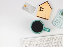 diseño plano de modelo de casa de madera, taza azul de café negro, calculadora azul, tarjeta de crédito y teclado de computadora sobre fondo blanco con espacio para copiar. concepto de compra de vivienda. foto