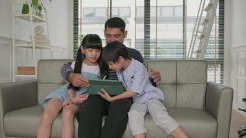 gelukkig Aziatisch Thais familie, pa, en kinderen hebben pret gebruik makend van digitaal tablet samen Aan sofa in de huis leven kamer, een lief vrije tijd weekend, en huiselijk welzijn levensstijl met internet technologie. video