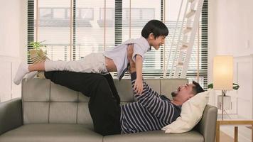 Asian Thai family together, father lies down and fun plays with son by lifting, spread arms like flying airplane on living room sofa, happy leisure times, lovely weekend, wellbeing domestic lifestyle.