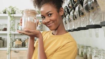 Portrait of young Black female customer smiles and looks at camera, shops with reusable glass jars in refill store, zero-waste grocery, and plastic-free, environment-friendly, sustainable lifestyles. video