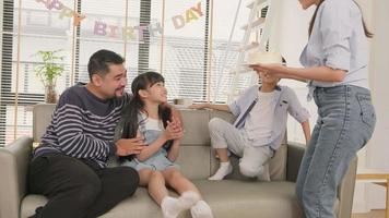 Happy Asian Thai family, young daughter is surprised with birthday cake, blows out candle, and cheerful celebrates party with parents together in living room, well-being domestic home event lifestyle.