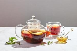 Fruit tea with apples and thyme and honey in glass teapot and cup on white background with hard shadows