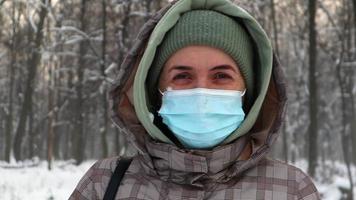 Lycklig kvinna i en skyddande medicinsk mask gläds i de snöfall utomhus i en stad parkera i vinter- mot de bakgrund av träd. positiv kvinna kvinna gläds i vinter- natur och vit snö. video