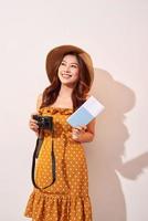 Portrait of a happy young woman in hat holding camera and showing passport while standing isolated over beige background photo