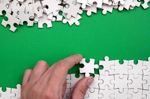The hand folds a white jigsaw puzzle and a pile of uncombed puzzle pieces lies against the background of the green surface. Texture photo with space for text