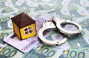 Small toy house and handcuffs is lies on a set of green monetary denominations of 100 euros. A lot of money forms an infinite heap photo